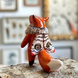 Könnte beinhalten: Eine glänzende Keramik-Fuchsfigur mit leuchtend orangefarbenem Körper und weißer Schwanzspitze. Der Fuchs hat einen strukturierten, graubraunen Kragen um den Hals und ein Paar Mottenflügel auf dem Rücken, die komplexe Muster mit gelben Augenflecken zeigen.