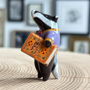Pode incluir: Uma pequena estatueta de cerâmica de um texugo vestindo uma camisa roxa com acabamento laranja. O texugo segura um livro retangular laranja com um padrão de videira verde e duas faces de abóbora, com o texto Pumpkin Patch na capa.