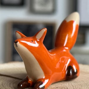 Puede incluir: Una figura de zorro de cerámica brillante con un cuerpo de color naranja vibrante, pecho y punta de la cola de color crema. El zorro tiene patas de color marrón oscuro, orejas puntiagudas y una nariz negra pintada, en posición sentada.