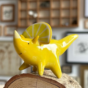 Könnte beinhalten: Eine glänzend gelbe Keramik-Dinosaurierfigur in Form eines Triceratops. Die Kreatur hat einen leuchtend gelben Körper mit einer glatten, reflektierenden Oberfläche und einen markanten Kopfschild, der an eine Zitronenscheibe erinnert.
