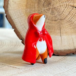 May include: A small ceramic fox figurine wearing a bright red hooded cape with a bow at the neck. The fox has a white chest and face with orange fur on its sides and back. The figurine is glossy and smooth, standing upright with black-tipped feet.