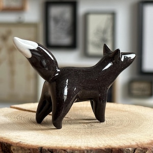 Puede incluir: Una pequeña figura de zorro de cerámica negra brillante con un acabado liso y reflectante. El zorro está representado en posición de pie con la cabeza ligeramente girada hacia la derecha y la cola apuntando hacia arriba, con una punta blanca.