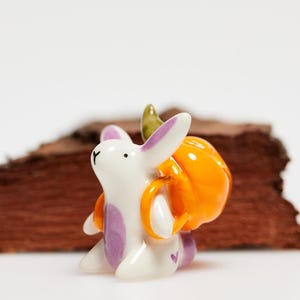 Könnte beinhalten: Eine kleine weiße Keramik-Hasenfigur mit violetten Akzenten an Ohren und Bauch, die einen leuchtend orangefarbenen Kürbis auf dem Rücken trägt. Der Hase hat ein winziges violettes Herzdetail an der Seite und einen kleinen grünen Stiel am Kürbis.