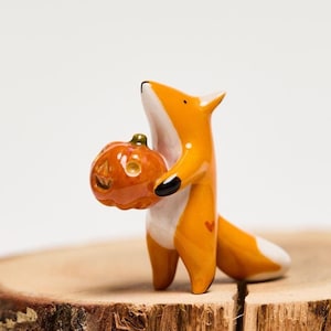 May include: A small, glossy ceramic fox figurine with orange and white fur, black-tipped ears, and a white-tipped tail. The fox is standing upright and holding a small, round, orange jack-o'-lantern pumpkin with carved facial features.