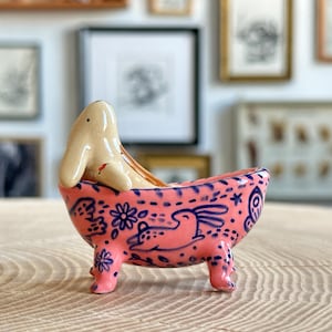Può includere: Una statuetta in ceramica di un coniglio beige seduto all'interno di una vasca da bagno rosa con quattro piedini. L'esterno della vasca è decorato con un motivo blu scuro raffigurante piccoli conigli, fiori e foglie. Il coniglio ha un segno rosso.