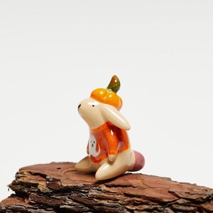 Może przedstawiać: Mała ceramiczna figurka beżowego królika w pomarańczowej czapce z dyni i pomarańczowym swetrze z białym nadrukiem duszka z przodu. Królik ma długie uszy, gładkie, błyszczące wykończenie oraz mały różowy ogonek i siedzi w wyprostowanej pozycji.