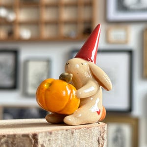 Könnte beinhalten: Eine Keramikfigur eines beigen Hasen mit einer hohen, glänzenden roten Zipfelmütze. Der Hase sitzt und hält einen leuchtend orangefarbenen Kürbis mit grünem Stiel. Ein kleines rotes Herz ist auf der Hüfte des Hasen in der Nähe des Schwanzes aufgemalt.