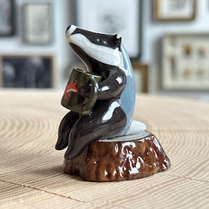 May include: A ceramic badger figurine with black and white fur markings, sitting on a brown textured tree stump base. The badger is holding a small olive green mug featuring a tiny orange fox illustration on its side, with a glossy finish.