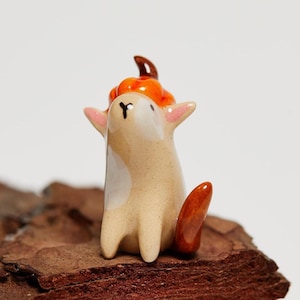 Peut inclure: Une petite figurine en céramique beige lisse représentant une créature ressemblant à un renard avec des oreilles pointues et une tache blanche sur la poitrine. La créature porte une citrouille orange brillante sur la tête et une queue orange foncé.