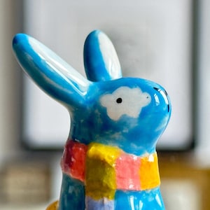 Op de afbeelding: Een keramieken konijnenbeeldje met een glanzende blauwe afwerking, voorzien van een witte wolkachtige vlek op de wang en een kleurrijke geblokte sjaal in tinten geel, roze, oranje en paars om de nek.