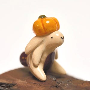 Könnte beinhalten: Eine kleine, cremefarbene Keramik-Hasenfigur mit glänzender Oberfläche. Der Hase hat einen dunkelbraunen Fleck auf der Brust und ein kleines, rundes, orangefarbenes kürbisförmiges Ornament auf dem Kopf. Er hat einfache, schwarz aufgemalte Augen und Nase.
