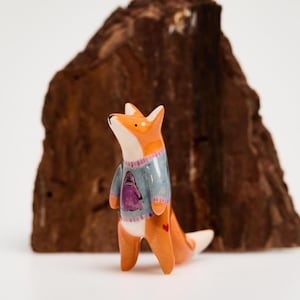 Puede incluir: Una pequeña figura de zorro de cerámica de pie, pintada en naranja y blanco con un suéter azul claro que presenta un estampado gráfico morado. El zorro tiene una cola con la punta blanca y un pequeño detalle de corazón rojo en el costado.