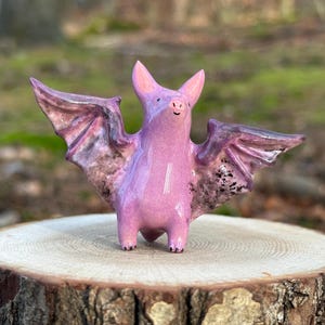May include: A small, glossy ceramic bat figurine in a vibrant shade of pink. The bat features pointed ears, a small snout, and outstretched wings with mottled dark purple and black detailing on the underside, standing on two short legs.