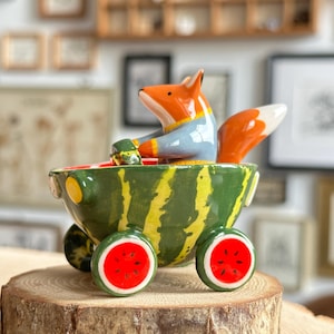 May include: A ceramic figurine of an orange and white fox wearing a blue sweater, sitting inside a green and yellow striped watermelon-shaped cart. The cart features four wheels designed to look like watermelon slices with red centers and black seeds.