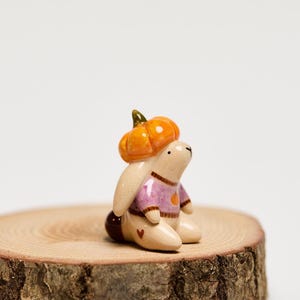 Könnte beinhalten: Eine kleine Keramikfigur eines cremefarbenen Hasen, der einen violetten Pullover mit einem kleinen orangefarbenen Kürbismotiv trägt. Der Hase hat einen leuchtend orangefarbenen Kürbis auf dem Kopf und ein winziges braunes Herz an der Seite.