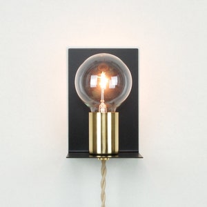 May include: A black metal wall sconce with a gold-colored light bulb socket and a clear glass light bulb. The light bulb is turned on and glowing. The sconce has a small shelf on the bottom.
