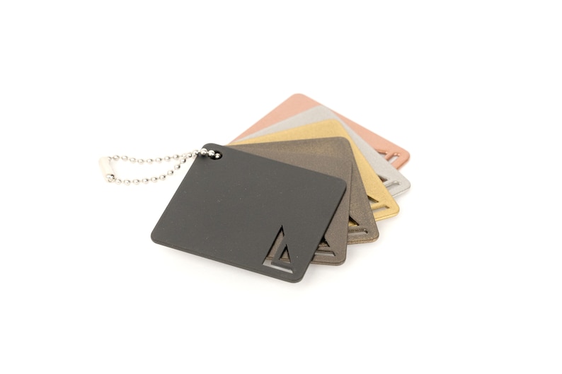 Op de afbeelding: Een set van vijf metalen kleurstalen bevestigd aan een zilveren ketting. De stalen zijn in tinten zwart, grijs, goud, koper en ros&eacute; goud.