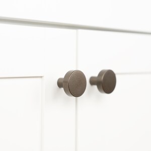 Op de afbeelding: Twee ronde, geborstelde nikkel kastknoppen zijn gemonteerd op een witte kastdeur.