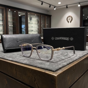 Puede incluir: Gafas de metal en tono dorado con montura cuadrada y bordes claros. Las varillas presentan detalles metálicos intrincados y el logotipo de Chrome Hearts es visible en la lente y en el estuche y la caja de cuero negro incluidos.
