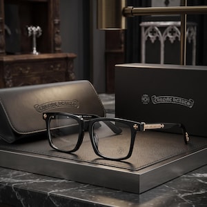 May include: Black rectangular spectacles with clear lenses and gold-coloured metal detailing on the temples and hinges. The frames feature small cross emblems. A matching black leather-effect case and a black box with the Chrome Hearts logo are included.