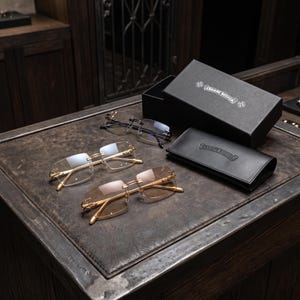 Puede incluir: Tres pares de gafas sin montura con patillas y puente de metal en tono dorado, con lentes transparentes y tintadas. El conjunto incluye una caja de almacenamiento rectangular negra y un estuche de piel sintética negra, ambos con el logo Chrome Hearts.