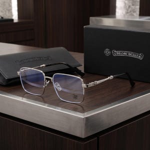 May include: Silver-toned square-framed spectacles featuring thin metal rims and black temple tips. The temples display intricate metallic detailing. A small cross emblem is etched onto the right lens. Includes a black case and box with the Chrome Hearts logo.