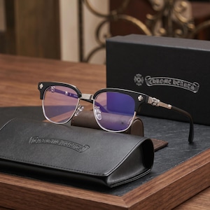 May include: Black and silver-toned browline glasses with blue-tinted lenses. The frames feature metallic cross emblems on the temples and bridge. The glasses are displayed with a black leather-textured case embossed with the Chrome Hearts logo.