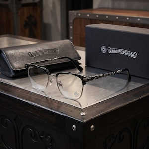 May include: Chrome Hearts spectacles featuring a black browline frame with silver-tone metal rims and temples. The temples are adorned with small silver cross motifs. A small cross logo is printed on the left lens. Includes a black case and box.