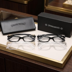 Puede incluir: Dos pares de gafas rectangulares, una negra y otra transparente, con detalles metálicos en forma de espada en las patillas. Las monturas se presentan junto a un estuche de cuero negro y una caja negra, ambos con el nombre Chrome Hearts grabado.