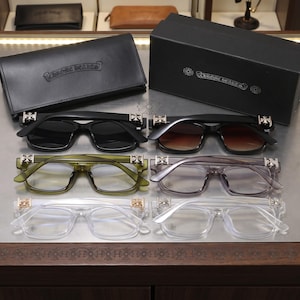 Può includere: Sei paia di occhiali da vista e da sole Chrome Hearts disposti su tre file. Le montature sono in acetato nero, sfumato marrone, verde oliva, grigio traslucido e trasparente, tutte con emblemi a croce color argento sulle aste.