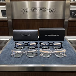 Op de afbeelding: Vier brillen van Chrome Hearts op een displayoppervlak. Twee paren hebben zwarte monturen en twee paren hebben transparante monturen. Alle vier de brillen hebben lichtgekleurde, getextureerde pootjes met metalen accenten. Een etui en doos zijn inbegrepen.