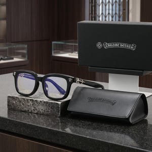 Puede incluir: Gafas de montura negra con lentes tintadas en azul y detalles plateados de flor de lis en las patillas. El conjunto incluye un estuche rígido de piel sintética negra y una caja de presentación negra, ambos con el texto Chrome Hearts.