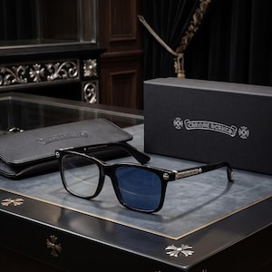 Pode incluir: Óculos de armação preta com detalhes em metal prateado nas hastes e dobradiças. As lentes apresentam um tom azulado. Um estojo com textura de couro preto e uma caixa preta com o texto prateado CHROME HEARTS estão posicionados atrás.