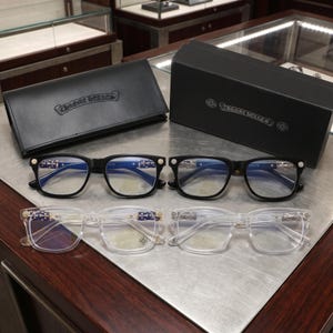 May include: Two pairs of black-framed glasses and two pairs of clear-framed glasses with metallic temple accents featuring cross motifs. Each pair includes a small circular logo on the lens. A black leather case and a black box both display the Chrome Hearts logo.