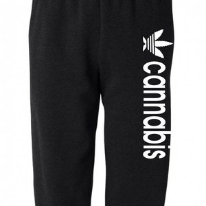 Op de afbeelding: Zwarte sweatpants met een wit cannabisbladlogo en het woord "cannabis" verticaal gedrukt op het rechterbeen.