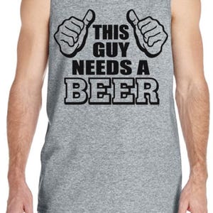 May include: Gray tank top with the text "THIS GUY NEEDS A BEER" in black block letters. The design includes two thumbs-up gestures. The shirt is a sleeveless style.
