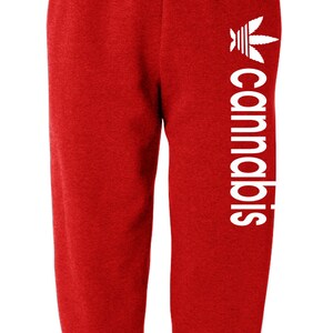 May include: Red sweatpants with the word "cannabis" printed vertically in white on the left leg. The design includes a white cannabis leaf logo. The pants have an elastic waistband and cuffs.