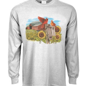 May include: A gray long-sleeve t-shirt with a red cardinal perched on a birdhouse. The birdhouse is surrounded by sunflowers and a red barn in the background.