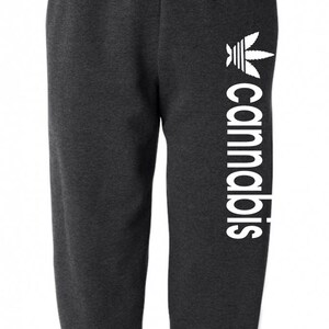 May include: Dark gray sweatpants with a white cannabis leaf logo and the word "cannabis" printed vertically. The sweatpants have an elastic waistband and cuffs, suitable for casual wear.