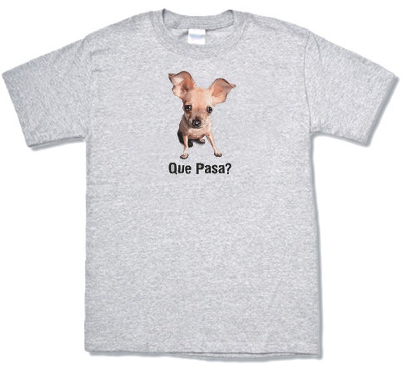 May include: A gray t-shirt with a brown and white chihuahua dog wearing a curious expression. The dog is looking directly at the viewer. The text "Que Pasa?" is printed below the dog.