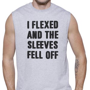 May include: A sleeveless gray t-shirt with the text "I Flexed and the Sleeves Fell Off" in black. The shirt is worn by a person wearing dark blue jeans.
