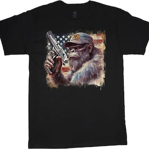 Può includere: Maglietta nera con una grafica di un gorilla che indossa un cappello con la scritta "RAD" e tiene una pistola davanti a una bandiera americana.