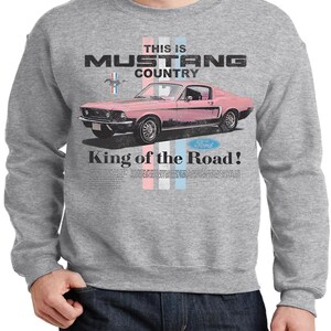 May include: A gray sweatshirt featuring a pink vintage Mustang car graphic. The text reads "THIS IS MUSTANG COUNTRY" and "King of the Road!" in a distressed font. The Ford logo is also visible.