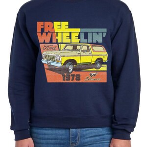 Puede incluir: Sudadera azul marino con un gráfico de un Ford Bronco amarillo de 1978. El diseño incluye las palabras "FREE WHEELIN'" en naranja, amarillo y azul, con el logotipo de Ford y la palabra "Bronco".