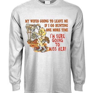 May include: A gray long-sleeve t-shirt with a cartoon illustration of a hunter holding a rifle. The text on the shirt reads: "MY WIFES GOING TO LEAVE ME IF I GO HUNTING ONE MORE TIME I'M SURE GOING TO MISS HER!"