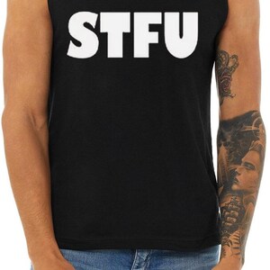 May include: Black sleeveless top with the white capital letters "STFU" printed across the front. The top is worn by a person wearing blue jeans. The person has a tattoo on their right arm.