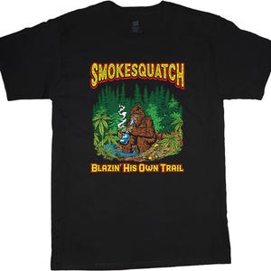 May include: Black t-shirt featuring a graphic of a brown Bigfoot smoking a pipe, with the text "SMOKESQUATCH" above and "BLAZIN' HIS OWN TRAIL" below. The design includes a forest scene with marijuana leaves.