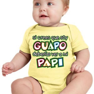 May include: A light yellow baby bodysuit with the Spanish text "si crees que soy GUAPO deberías ver a mi PAPI" in colorful lettering. The baby is fair-skinned with light brown hair and blue eyes.