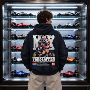 Könnte beinhalten: Ein dunkelblauer Kapuzenpullover mit einer großen Grafik auf der Rückseite, die den Formel-1-Fahrer Max Verstappen mit Rennhelm zeigt. Der Text lautet Formula 1 Driver Max Verstappen, ergänzt durch eine niederländische Flagge und die Zahl 01.
