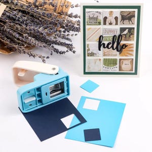 May include: A light blue square paper punch tool with a white handle, positioned next to a greeting card featuring a grid of nine small square images and the black script text hello, along with dark blue and light blue paper scraps and cutouts.